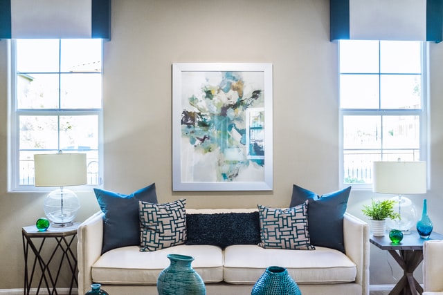 A white sofa between two windows with blue pillows and a painting above it