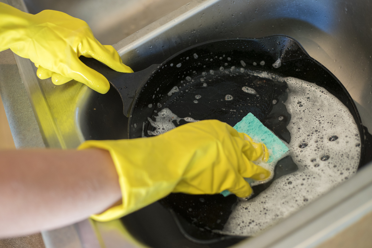 Hands of a woman cleaning a cast iron skillet using a sponge to remove the dirt